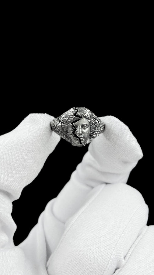 A high-contrast macro photograph of a heavy sterling silver ring resting against a pitch-black background. The ring features a three-dimensional sculpture of a classical Greek goddess's face, likely Athena. A jagged, deep crack splits the face vertically. The right side shows smooth, classical features, while the left side transitions into a rough, eroded, and porous texture resembling crumbling ancient stone. The silver is heavily oxidized t (etsy image id: 7831406732)