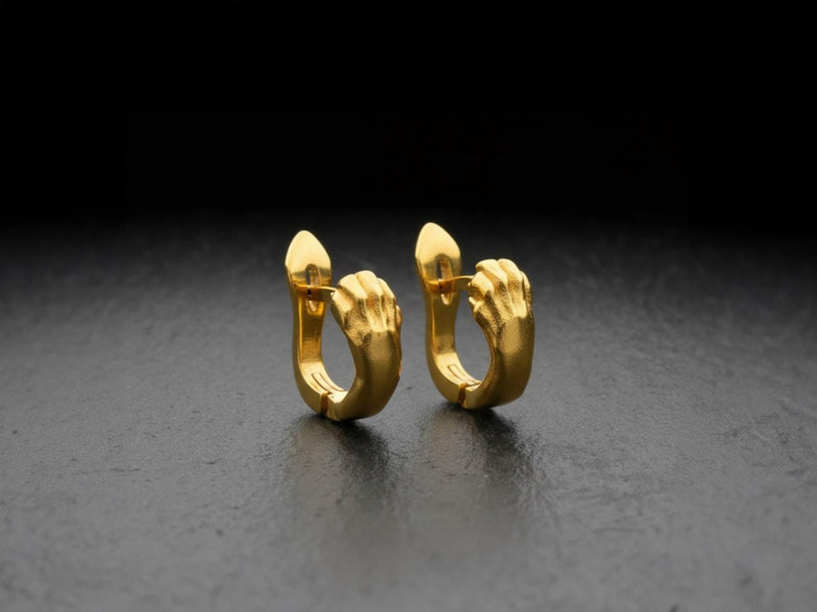 A close-up studio shot of a pair of sterling silver huggie hoop earrings resting on a dark slate surface. The front of each earring is masterfully sculpted to resemble a realistic cat paw, complete with distinct knuckles, claws, and soft paw pads. The earrings feature a hinged mechanism that allows the paw to wrap seamlessly around the earlobe. The metal is treated with heavy black oxidation in the recesses, contrasting beautifully with the brigh (etsy image id: 4850613175)
