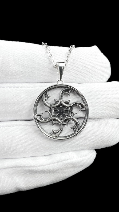 A detailed close-up of a circular sterling silver pendant resting on a dark blue, textured silk fabric. The pendant features an openwork design with swirling, crescent-like shapes radiating from a central four-pointed star. The intricate pattern resembles a gothic rose window or a celestial wheel. The raised metal surfaces are polished to a bright silver shine, contrasting beautifully against the dark, oxidized background recesses. (etsy image id: 7830974908)