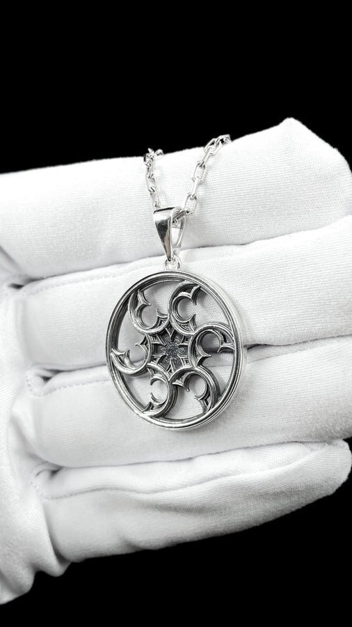 A detailed close-up of a circular sterling silver pendant resting on a dark blue, textured silk fabric. The pendant features an openwork design with swirling, crescent-like shapes radiating from a central four-pointed star. The intricate pattern resembles a gothic rose window or a celestial wheel. The raised metal surfaces are polished to a bright silver shine, contrasting beautifully against the dark, oxidized background recesses. (etsy image id: 7830974908)