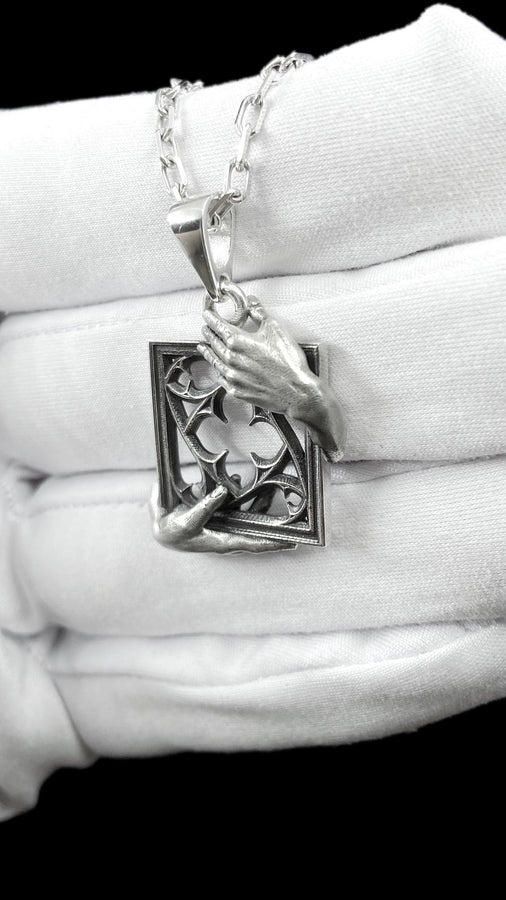 A close-up studio photograph of a highly detailed sterling silver pendant resting on a luxurious, dark blue silk fabric with silvery thread patterns. The pendant's striking design features two realistically sculpted, three-dimensional hands emerging from the sides to gently grasp a rectangular Gothic window frame. Inside the frame sits intricate openwork tracery, forming a classic quatrefoil motif. The polished silver highlights contrast with (etsy image id: 7879251833)