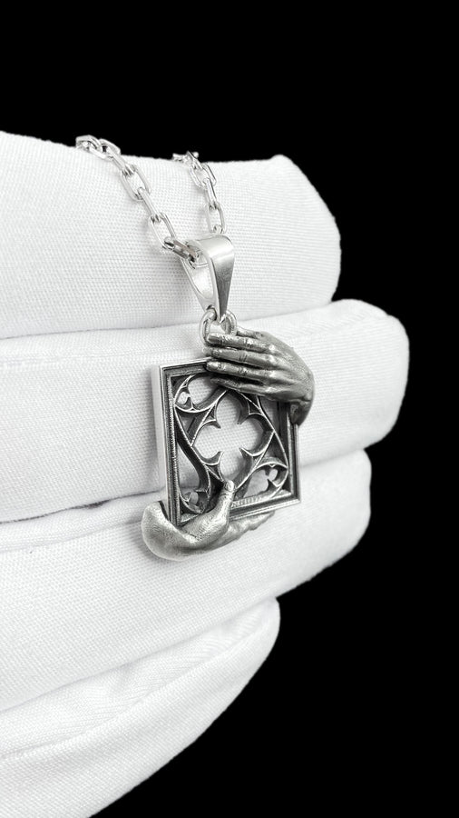 A close-up studio photograph of a highly detailed sterling silver pendant resting on a luxurious, dark blue silk fabric with silvery thread patterns. The pendant's striking design features two realistically sculpted, three-dimensional hands emerging from the sides to gently grasp a rectangular Gothic window frame. Inside the frame sits intricate openwork tracery, forming a classic quatrefoil motif. The polished silver highlights contrast with (etsy image id: 7879251833)