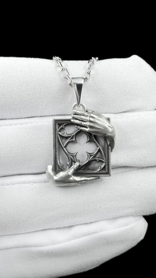 A close-up studio photograph of a highly detailed sterling silver pendant resting on a luxurious, dark blue silk fabric with silvery thread patterns. The pendant's striking design features two realistically sculpted, three-dimensional hands emerging from the sides to gently grasp a rectangular Gothic window frame. Inside the frame sits intricate openwork tracery, forming a classic quatrefoil motif. The polished silver highlights contrast with (etsy image id: 7879251833)