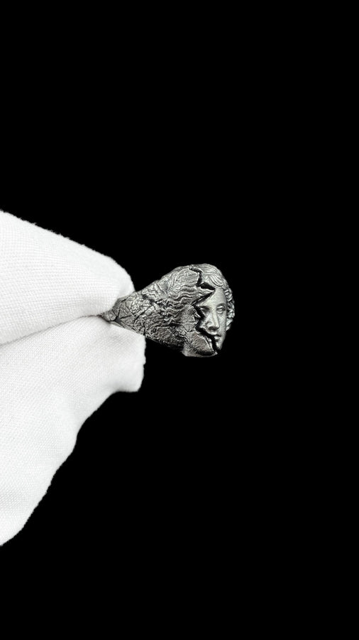A high-contrast macro photograph of a heavy sterling silver ring resting against a pitch-black background. The ring features a three-dimensional sculpture of a classical Greek goddess's face, likely Athena. A jagged, deep crack splits the face vertically. The right side shows smooth, classical features, while the left side transitions into a rough, eroded, and porous texture resembling crumbling ancient stone. The silver is heavily oxidized t (etsy image id: 7831406732)