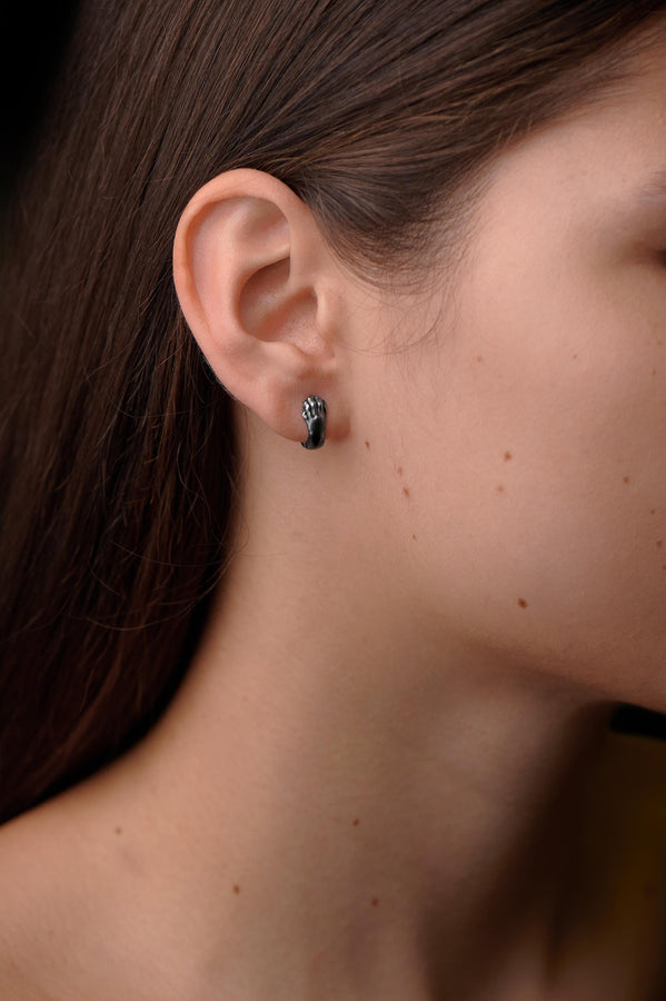 A close-up studio shot of a pair of sterling silver huggie hoop earrings resting on a dark slate surface. The front of each earring is masterfully sculpted to resemble a realistic cat paw, complete with distinct knuckles, claws, and soft paw pads. The earrings feature a hinged mechanism that allows the paw to wrap seamlessly around the earlobe. The metal is treated with heavy black oxidation in the recesses, contrasting beautifully with the brigh (etsy image id: 4850613175)