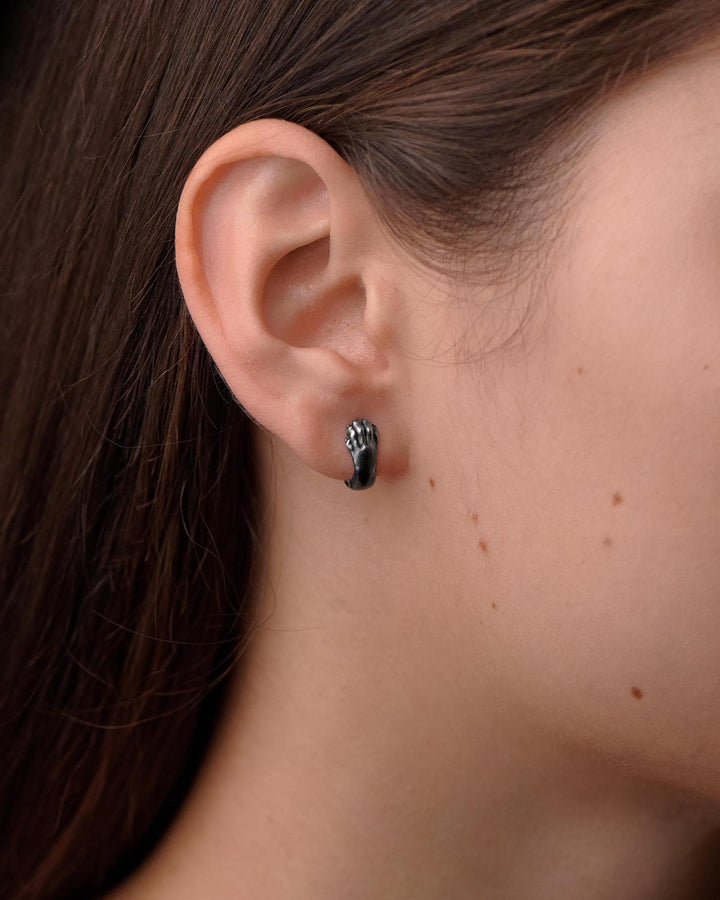 A close-up studio shot of a pair of sterling silver huggie hoop earrings resting on a dark slate surface. The front of each earring is masterfully sculpted to resemble a realistic cat paw, complete with distinct knuckles, claws, and soft paw pads. The earrings feature a hinged mechanism that allows the paw to wrap seamlessly around the earlobe. The metal is treated with heavy black oxidation in the recesses, contrasting beautifully with the brigh (etsy image id: 4850613175)