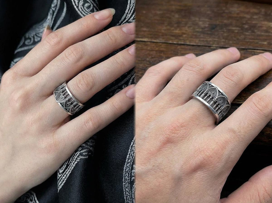 A close-up of a wide sterling silver ring resting on dark patterned fabric. The band features a continuous arcade of pointed gothic arches supported by slender columns, with small cross motifs in the spandrels. The silver is heavily oxidized in the recesses to highlight the raised architectural details. The intricate relief mimics the stone tracery found in medieval cathedral windows, offering a rugged yet highly sophisticated and historically ac (etsy image id: 7958165023)