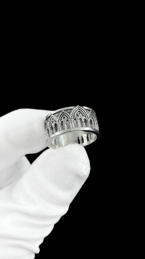 A close-up of a wide sterling silver ring resting on dark patterned fabric. The band features a continuous arcade of pointed gothic arches supported by slender columns, with small cross motifs in the spandrels. The silver is heavily oxidized in the recesses to highlight the raised architectural details. The intricate relief mimics the stone tracery found in medieval cathedral windows, offering a rugged yet highly sophisticated and historically ac (etsy image id: 7958165023)