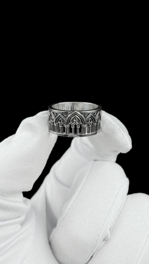 A close-up of a wide sterling silver ring resting on dark patterned fabric. The band features a continuous arcade of pointed gothic arches supported by slender columns, with small cross motifs in the spandrels. The silver is heavily oxidized in the recesses to highlight the raised architectural details. The intricate relief mimics the stone tracery found in medieval cathedral windows, offering a rugged yet highly sophisticated and historically ac (etsy image id: 7958165023)
