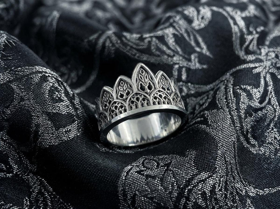 A close-up studio shot of a wide sterling silver ring resting on a dark patterned brocade fabric. The band features a majestic crown or tiara silhouette made of continuous pointed gothic arches. Inside each arch is delicate, openwork filigree that resembles the ornamental stone latticework of cathedral windows. The silver is oxidized in the recesses to highlight the raised, polished metal. (etsy image id: 7910304990)