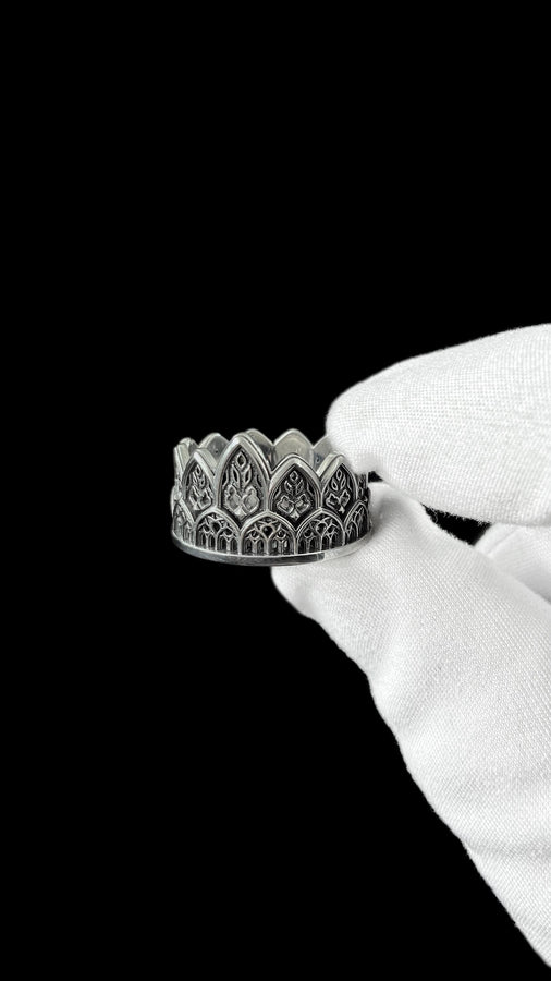 A close-up studio shot of a wide sterling silver ring resting on a dark patterned brocade fabric. The band features a majestic crown or tiara silhouette made of continuous pointed gothic arches. Inside each arch is delicate, openwork filigree that resembles the ornamental stone latticework of cathedral windows. The silver is oxidized in the recesses to highlight the raised, polished metal. (etsy image id: 7910304990)