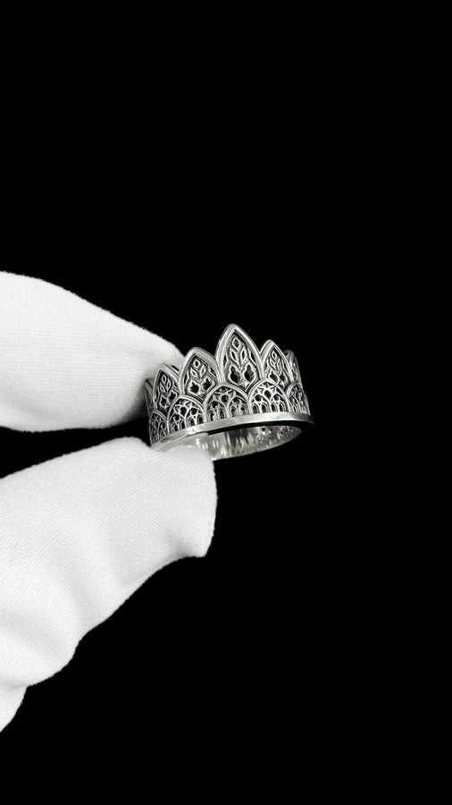 A close-up studio shot of a wide sterling silver ring resting on a dark patterned brocade fabric. The band features a majestic crown or tiara silhouette made of continuous pointed gothic arches. Inside each arch is delicate, openwork filigree that resembles the ornamental stone latticework of cathedral windows. The silver is oxidized in the recesses to highlight the raised, polished metal. (etsy image id: 7910304990)