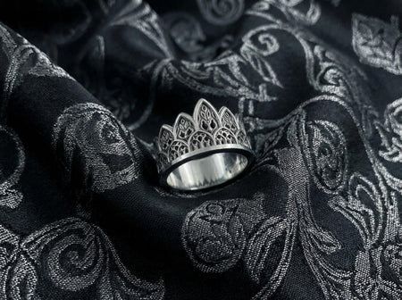 A close-up studio shot of a wide sterling silver ring resting on a dark patterned brocade fabric. The band features a majestic crown or tiara silhouette made of continuous pointed gothic arches. Inside each arch is delicate, openwork filigree that resembles the ornamental stone latticework of cathedral windows. The silver is oxidized in the recesses to highlight the raised, polished metal. (etsy image id: 7910304990)