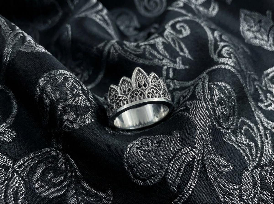 A close-up studio shot of a wide sterling silver ring resting on a dark patterned brocade fabric. The band features a majestic crown or tiara silhouette made of continuous pointed gothic arches. Inside each arch is delicate, openwork filigree that resembles the ornamental stone latticework of cathedral windows. The silver is oxidized in the recesses to highlight the raised, polished metal. (etsy image id: 7910304990)