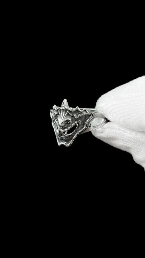 A close-up studio shot of a sterling silver ring featuring a detailed Japanese Oni mask. The demon face has sharp fangs, a wide menacing grin, and two distinct horns protruding from its forehead. The band features openwork cutouts on the sides, merging seamlessly with the mask. The metal is heavily oxidized to a dark charcoal in the recesses, making the highly polished silver ridges of the fierce expression pop against the dark reflective backgro (etsy image id: 7959798327)