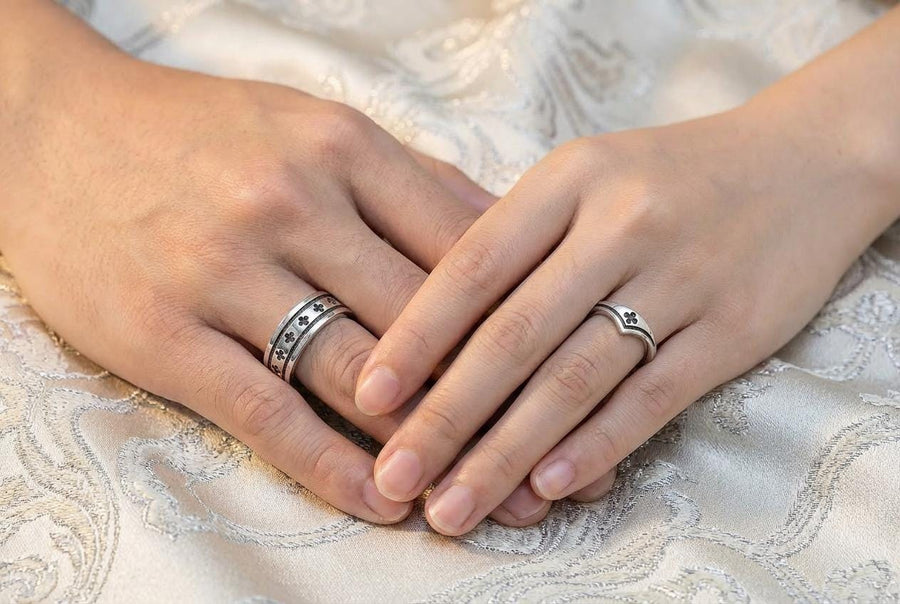 A high-resolution studio shot of a matching sterling silver wedding ring set resting on a sparkling, icy white surface. The set includes a wide unisex band and a delicate, V-shaped chevron ring. Both feature polished medieval quatrefoil motifs—four-lobed geometric shapes—set against a deeply textured, oxidized dark grey background that resembles crushed stone or stippled concrete.
