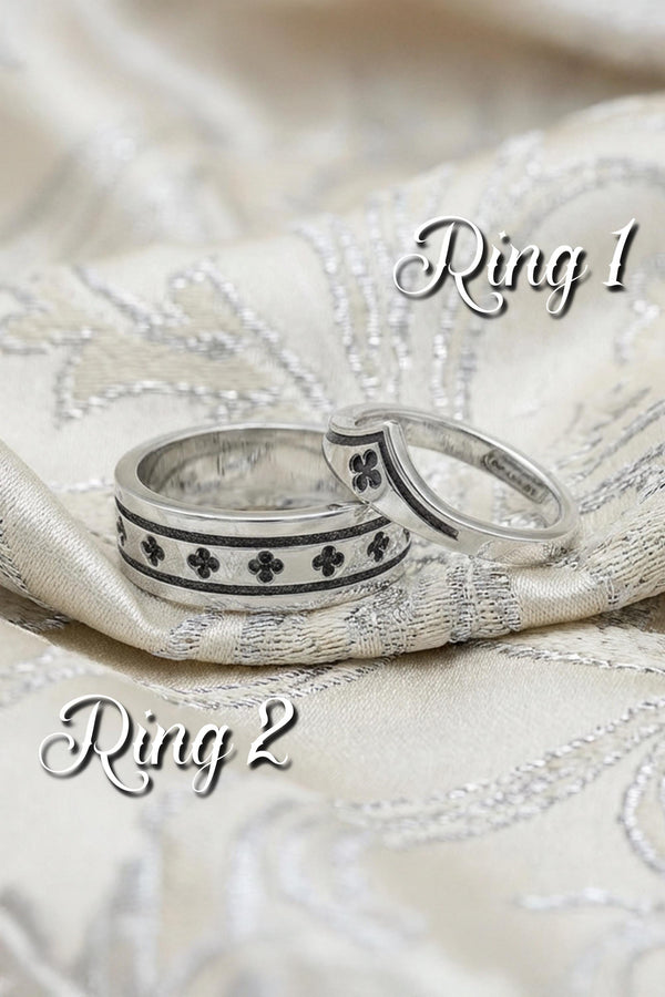 A high-resolution studio shot of a matching sterling silver wedding ring set resting on a sparkling, icy white surface. The set includes a wide unisex band and a delicate, V-shaped chevron ring. Both feature polished medieval quatrefoil motifs—four-lobed geometric shapes—set against a deeply textured, oxidized dark grey background that resembles crushed stone or stippled concrete.