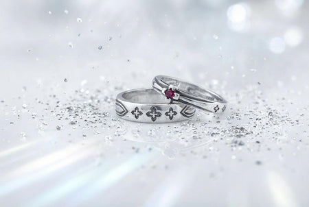 A high-resolution studio shot of a matching sterling silver gothic wedding ring set resting on a sparkling, white surface scattered with tiny crystals. The beautiful set includes a wide men's band and a narrower engagement ring centering a vibrant round red ruby. Both rings feature precisely polished medieval quatrefoil motifs four-lobed geometric shapes set against a deeply oxidized, stippled dark grey background, beautifully highlighting th