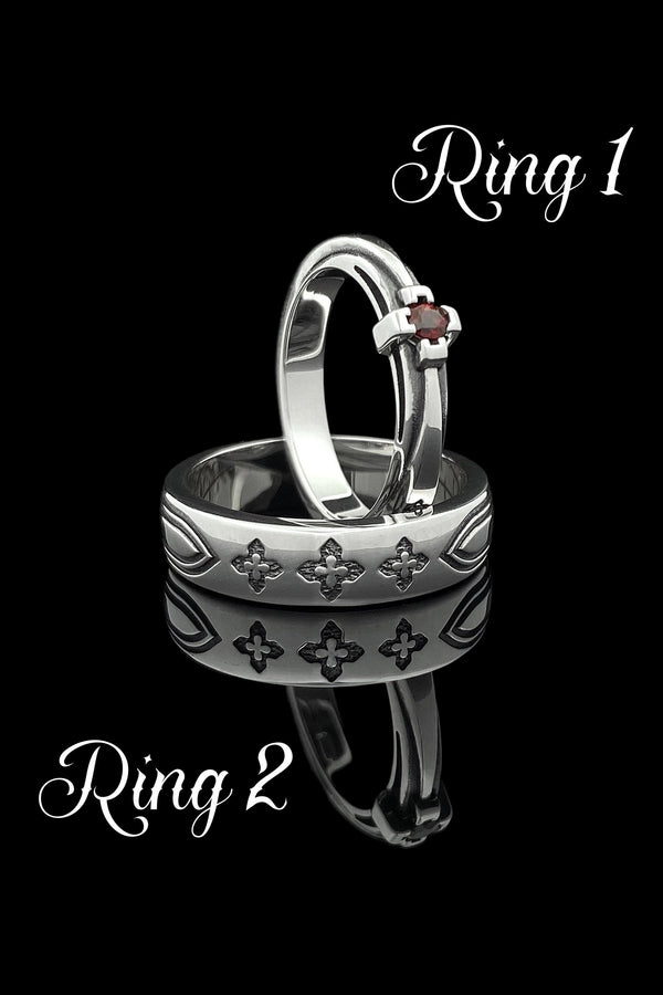 A high-resolution studio shot of a matching sterling silver gothic wedding ring set resting on a sparkling, white surface scattered with tiny crystals. The beautiful set includes a wide men's band and a narrower engagement ring centering a vibrant round red ruby. Both rings feature precisely polished medieval quatrefoil motifs four-lobed geometric shapes set against a deeply oxidized, stippled dark grey background, beautifully highlighting th