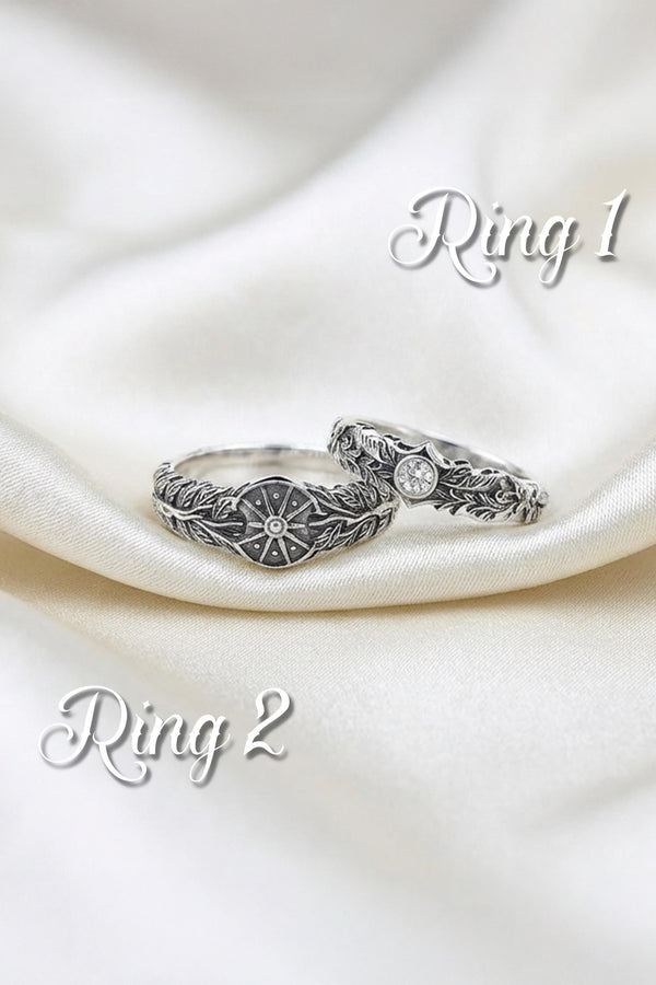 A macro studio shot of two matching antique-style sterling silver wedding rings resting on a sparkling, icy white surface. The wider men's band features a circular sunburst or shield motif with radiating lines. The narrower engagement ring features a matching sunburst centering a round white gemstone. Both bands are richly decorated with raised leaf and vine engravings, set against a dark oxidized background.