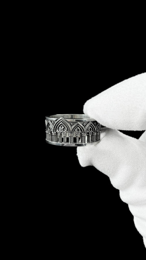 A close-up of a wide sterling silver ring resting on dark patterned fabric. The band features a continuous arcade of pointed gothic arches supported by slender columns, with small cross motifs in the spandrels. The silver is heavily oxidized in the recesses to highlight the raised architectural details. The intricate relief mimics the stone tracery found in medieval cathedral windows, offering a rugged yet highly sophisticated and historically ac (etsy image id: 7958165023)