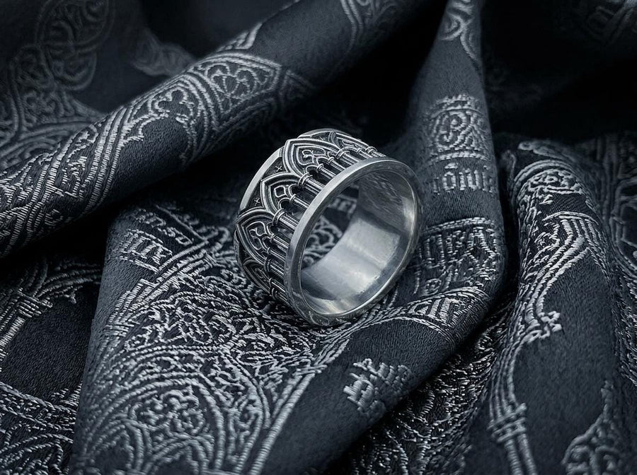 A close-up of a wide sterling silver ring resting on dark patterned fabric. The band features a continuous arcade of pointed gothic arches supported by slender columns, with small cross motifs in the spandrels. The silver is heavily oxidized in the recesses to highlight the raised architectural details. The intricate relief mimics the stone tracery found in medieval cathedral windows, offering a rugged yet highly sophisticated and historically ac (etsy image id: 7958165023)