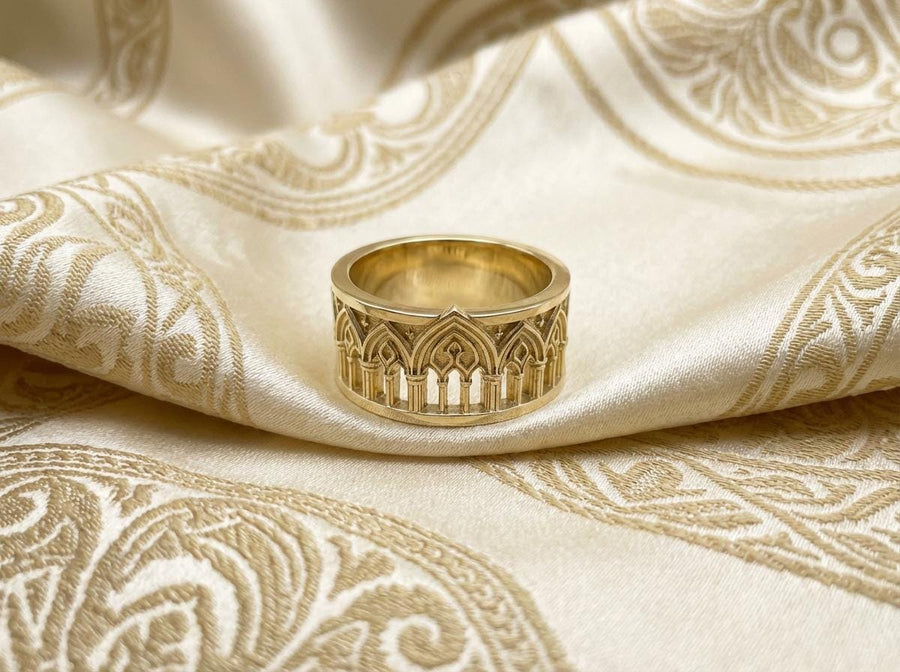 A close-up of a wide sterling silver ring resting on dark patterned fabric. The band features a continuous arcade of pointed gothic arches supported by slender columns, with small cross motifs in the spandrels. The silver is heavily oxidized in the recesses to highlight the raised architectural details. The intricate relief mimics the stone tracery found in medieval cathedral windows, offering a rugged yet highly sophisticated and historically ac (etsy image id: 7958165023)