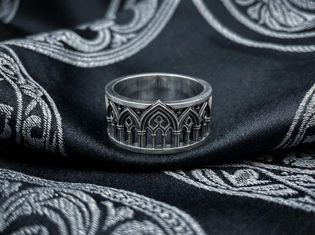 A close-up of a wide sterling silver ring resting on dark patterned fabric. The band features a continuous arcade of pointed gothic arches supported by slender columns, with small cross motifs in the spandrels. The silver is heavily oxidized in the recesses to highlight the raised architectural details. The intricate relief mimics the stone tracery found in medieval cathedral windows, offering a rugged yet highly sophisticated and historically ac (etsy image id: 7958165023)