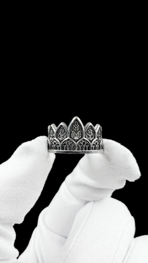 A close-up studio shot of a wide sterling silver ring resting on a dark patterned brocade fabric. The band features a majestic crown or tiara silhouette made of continuous pointed gothic arches. Inside each arch is delicate, openwork filigree that resembles the ornamental stone latticework of cathedral windows. The silver is oxidized in the recesses to highlight the raised, polished metal. (etsy image id: 7910304990)
