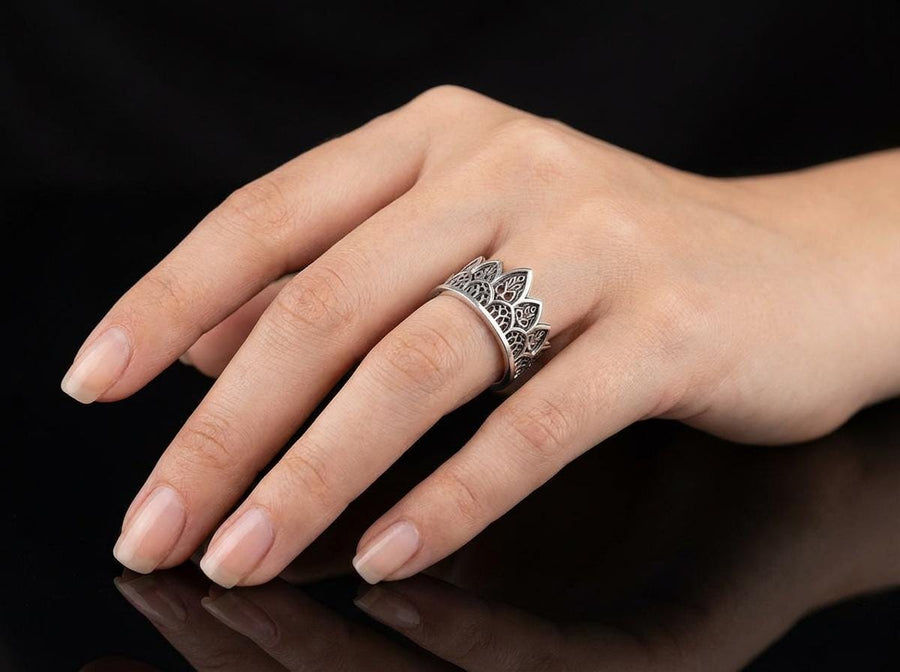 A close-up studio shot of a wide sterling silver ring resting on a dark patterned brocade fabric. The band features a majestic crown or tiara silhouette made of continuous pointed gothic arches. Inside each arch is delicate, openwork filigree that resembles the ornamental stone latticework of cathedral windows. The silver is oxidized in the recesses to highlight the raised, polished metal. (etsy image id: 7910304990)