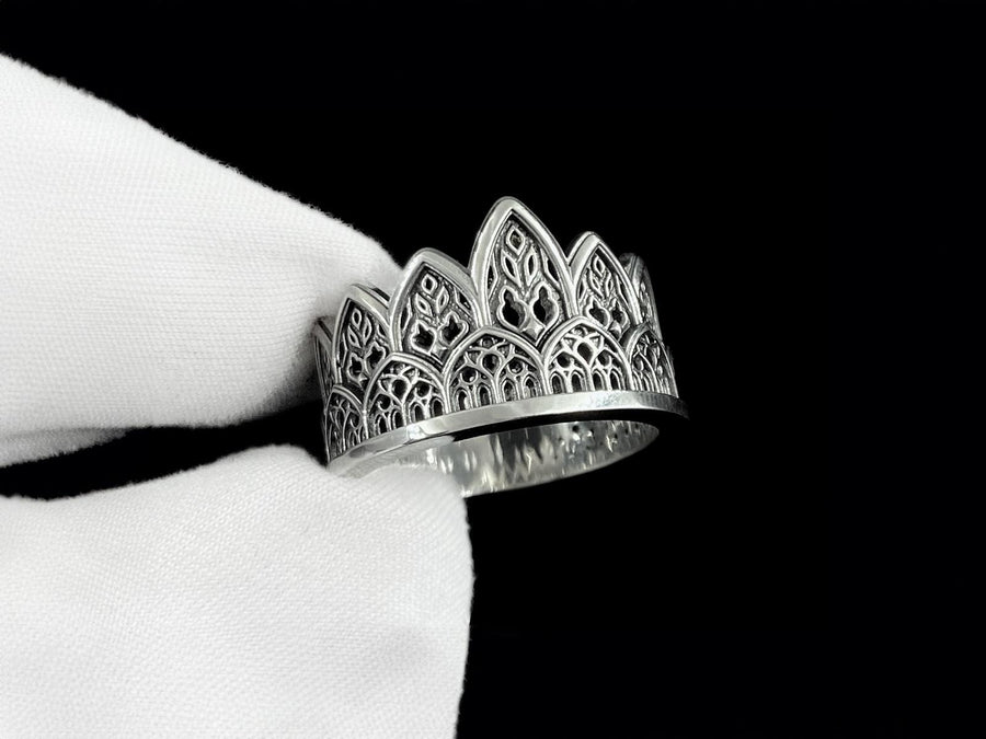 A close-up studio shot of a wide sterling silver ring resting on a dark patterned brocade fabric. The band features a majestic crown or tiara silhouette made of continuous pointed gothic arches. Inside each arch is delicate, openwork filigree that resembles the ornamental stone latticework of cathedral windows. The silver is oxidized in the recesses to highlight the raised, polished metal. (etsy image id: 7910304990)