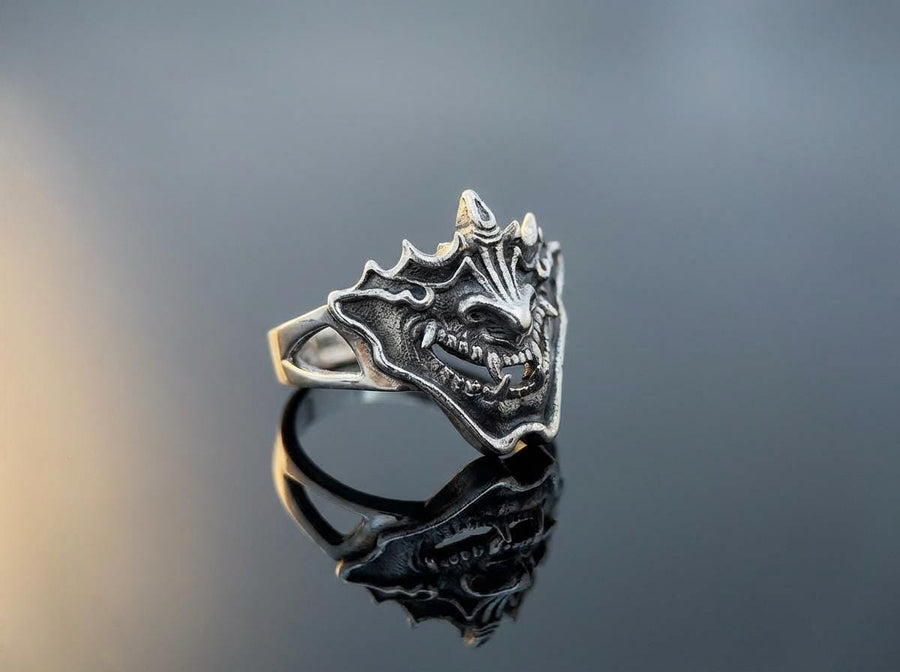 A close-up studio shot of a sterling silver ring featuring a detailed Japanese Oni mask. The demon face has sharp fangs, a wide menacing grin, and two distinct horns protruding from its forehead. The band features openwork cutouts on the sides, merging seamlessly with the mask. The metal is heavily oxidized to a dark charcoal in the recesses, making the highly polished silver ridges of the fierce expression pop against the dark reflective backgro (etsy image id: 7959798327)