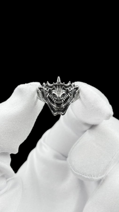 A close-up studio shot of a sterling silver ring featuring a detailed Japanese Oni mask. The demon face has sharp fangs, a wide menacing grin, and two distinct horns protruding from its forehead. The band features openwork cutouts on the sides, merging seamlessly with the mask. The metal is heavily oxidized to a dark charcoal in the recesses, making the highly polished silver ridges of the fierce expression pop against the dark reflective backgro (etsy image id: 7959798327)