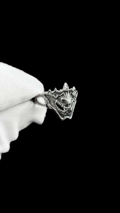A close-up studio shot of a sterling silver ring featuring a detailed Japanese Oni mask. The demon face has sharp fangs, a wide menacing grin, and two distinct horns protruding from its forehead. The band features openwork cutouts on the sides, merging seamlessly with the mask. The metal is heavily oxidized to a dark charcoal in the recesses, making the highly polished silver ridges of the fierce expression pop against the dark reflective backgro (etsy image id: 7959798327)