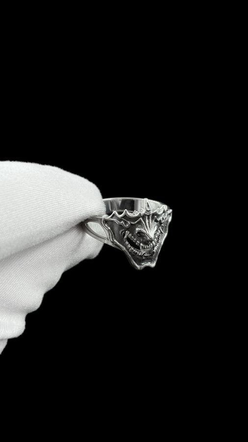 A close-up studio shot of a sterling silver ring featuring a detailed Japanese Oni mask. The demon face has sharp fangs, a wide menacing grin, and two distinct horns protruding from its forehead. The band features openwork cutouts on the sides, merging seamlessly with the mask. The metal is heavily oxidized to a dark charcoal in the recesses, making the highly polished silver ridges of the fierce expression pop against the dark reflective backgro (etsy image id: 7959798327)