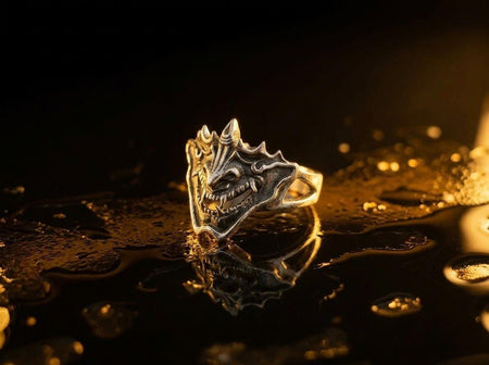 A close-up studio shot of a sterling silver ring featuring a detailed Japanese Oni mask. The demon face has sharp fangs, a wide menacing grin, and two distinct horns protruding from its forehead. The band features openwork cutouts on the sides, merging seamlessly with the mask. The metal is heavily oxidized to a dark charcoal in the recesses, making the highly polished silver ridges of the fierce expression pop against the dark reflective backgro (etsy image id: 7959798327)