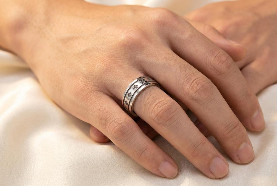 A high-resolution studio shot of a matching sterling silver wedding ring set resting on a sparkling, icy white surface. The set includes a wide unisex band and a delicate, V-shaped chevron ring. Both feature polished medieval quatrefoil motifs—four-lobed geometric shapes—set against a deeply textured, oxidized dark grey background that resembles crushed stone or stippled concrete.