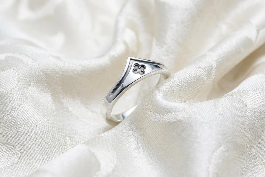 A high-resolution studio shot of a matching sterling silver wedding ring set resting on a sparkling, icy white surface. The set includes a wide unisex band and a delicate, V-shaped chevron ring. Both feature polished medieval quatrefoil motifs—four-lobed geometric shapes—set against a deeply textured, oxidized dark grey background that resembles crushed stone or stippled concrete.