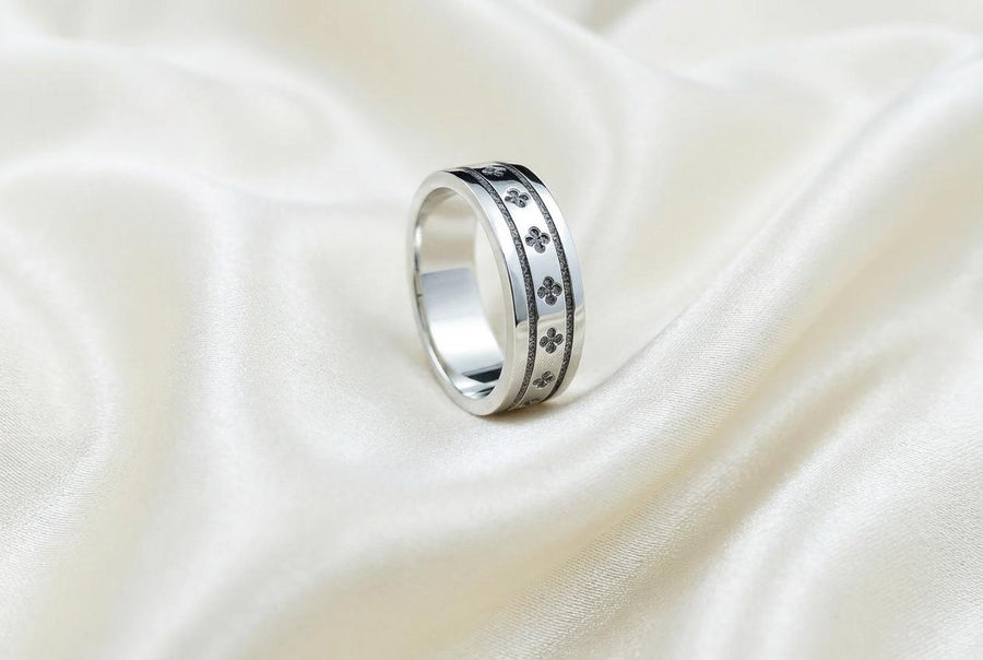 A high-resolution studio shot of a matching sterling silver wedding ring set resting on a sparkling, icy white surface. The set includes a wide unisex band and a delicate, V-shaped chevron ring. Both feature polished medieval quatrefoil motifs—four-lobed geometric shapes—set against a deeply textured, oxidized dark grey background that resembles crushed stone or stippled concrete.