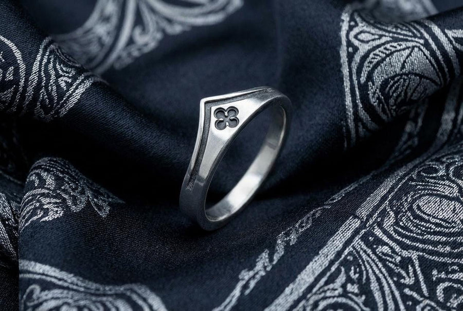 A high-resolution studio shot of a matching sterling silver wedding ring set resting on a sparkling, icy white surface. The set includes a wide unisex band and a delicate, V-shaped chevron ring. Both feature polished medieval quatrefoil motifs—four-lobed geometric shapes—set against a deeply textured, oxidized dark grey background that resembles crushed stone or stippled concrete.