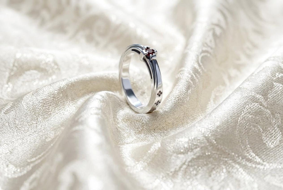 A high-resolution studio shot of a matching sterling silver gothic wedding ring set resting on a sparkling, white surface scattered with tiny crystals. The beautiful set includes a wide men's band and a narrower engagement ring centering a vibrant round red ruby. Both rings feature precisely polished medieval quatrefoil motifs four-lobed geometric shapes set against a deeply oxidized, stippled dark grey background, beautifully highlighting th
