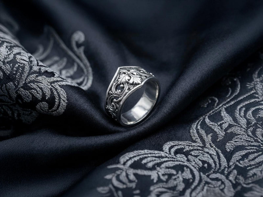A close-up studio photograph of a matching sterling silver gothic wedding ring set resting on a highly reflective black surface. The set includes a wide band and a pointed, V-shaped chevron ring. Both bands feature deeply carved, raised leaf and vine scrollwork. The engagement ring centers a round, dark red garnet gemstone. The silver is heavily oxidized, creating a stark contrast between the bright ridges and the dark recesses.
