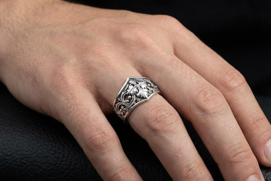 A close-up studio photograph of a matching sterling silver gothic wedding ring set resting on a highly reflective black surface. The set includes a wide band and a pointed, V-shaped chevron ring. Both bands feature deeply carved, raised leaf and vine scrollwork. The engagement ring centers a round, dark red garnet gemstone. The silver is heavily oxidized, creating a stark contrast between the bright ridges and the dark recesses.
