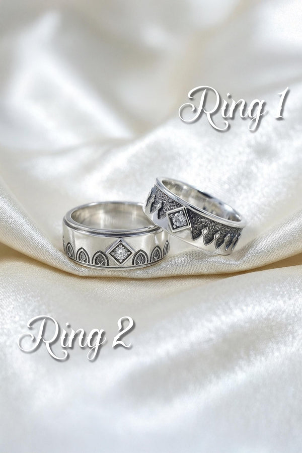 A high-resolution macro photograph of a matching sterling silver gothic wedding ring set resting on softly draped, luxurious white satin. The set features a wider masculine band and a slightly narrower feminine band. Both rings display a continuous engraved arcade of pointed medieval arches along the bottom edge, centering on a polished geometric rhombus frame that holds a brilliant round white gemstone. The polished silver beautifully contrasts