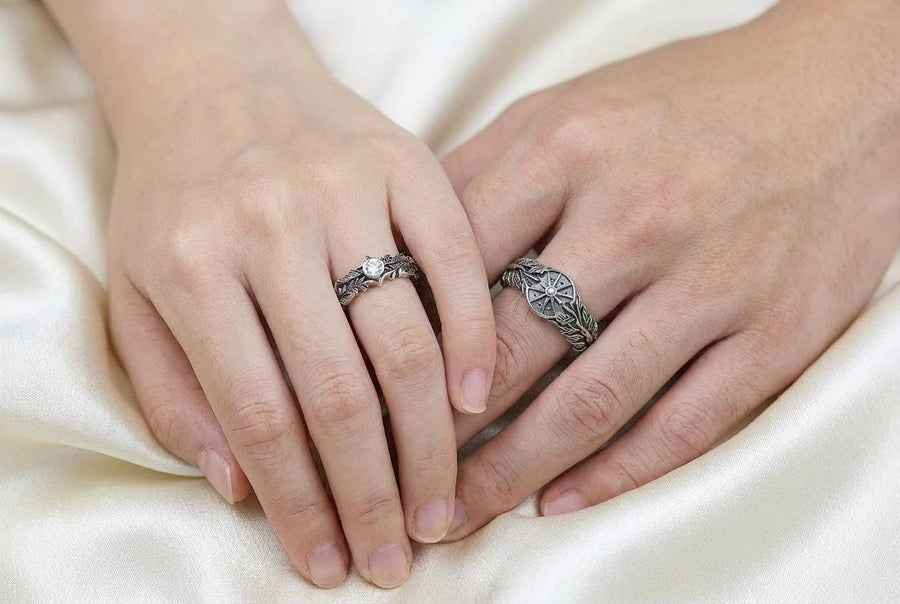 A macro studio shot of two matching antique-style sterling silver wedding rings resting on a sparkling, icy white surface. The wider men's band features a circular sunburst or shield motif with radiating lines. The narrower engagement ring features a matching sunburst centering a round white gemstone. Both bands are richly decorated with raised leaf and vine engravings, set against a dark oxidized background.