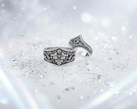 A close-up studio photograph of a matching sterling silver gothic wedding ring set resting on a highly reflective black surface. The set includes a wide band and a pointed, V-shaped chevron ring. Both bands feature deeply carved, raised leaf and vine scrollwork. The engagement ring centers a round, dark red garnet gemstone. The silver is heavily oxidized, creating a stark contrast between the bright ridges and the dark recesses.