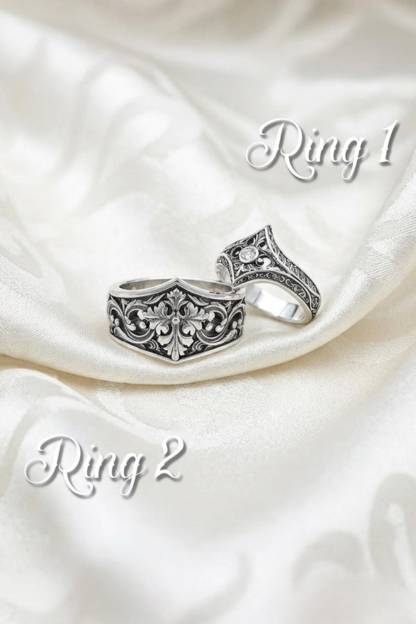 A close-up studio photograph of a matching sterling silver gothic wedding ring set resting on a highly reflective black surface. The set includes a wide band and a pointed, V-shaped chevron ring. Both bands feature deeply carved, raised leaf and vine scrollwork. The engagement ring centers a round, dark red garnet gemstone. The silver is heavily oxidized, creating a stark contrast between the bright ridges and the dark recesses.