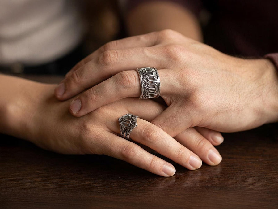 A high-resolution macro shot of a wide sterling silver wedding band resting on a sparkling, icy white surface. The ring features an intricate openwork design mimicking a Gothic cathedral window, complete with pointed lancet arches and delicate stone tracery. The polished silver highlights contrast beautifully against the dark, oxidized background, emphasizing the architectural depth of the piece.