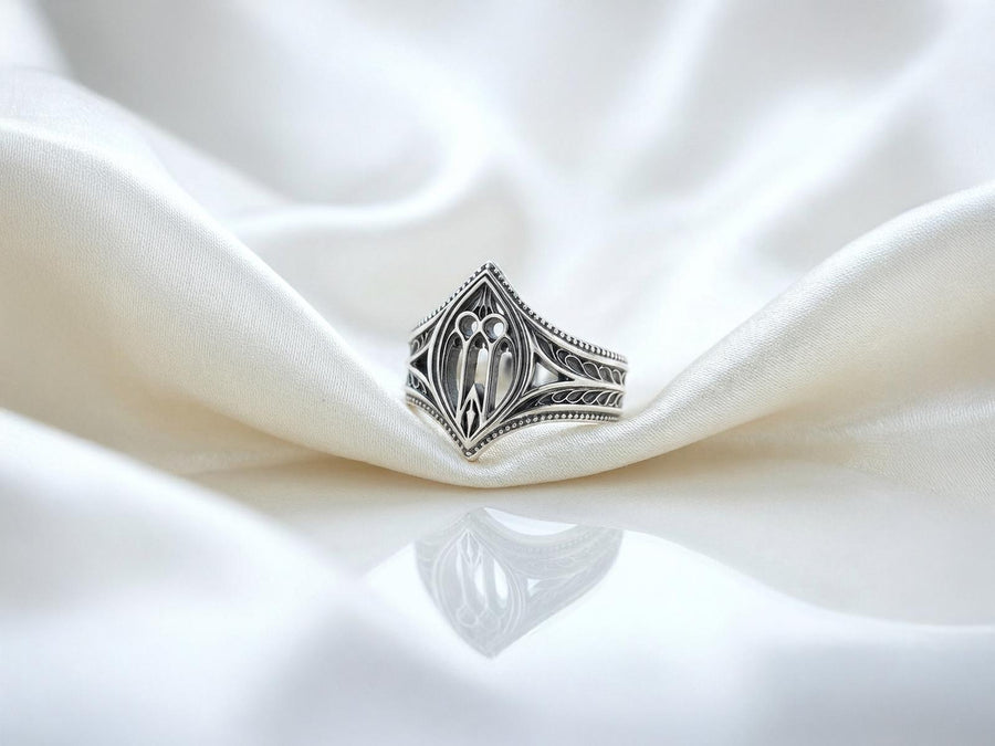 A high-resolution macro shot of a wide sterling silver wedding band resting on a sparkling, icy white surface. The ring features an intricate openwork design mimicking a Gothic cathedral window, complete with pointed lancet arches and delicate stone tracery. The polished silver highlights contrast beautifully against the dark, oxidized background, emphasizing the architectural depth of the piece.