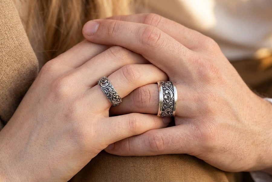 A close-up studio shot of a matching organic gothic wedding band set in sterling silver, resting on a dark, reflective surface. The rings feature a continuous, high-relief pattern of intricate sterling silver vine patterns and swirling leaves. The recessed areas are heavily oxidized to a dark matte black, creating a high-contrast shadow effect that makes the polished organic scrollwork stand out brilliantly against the dark background.